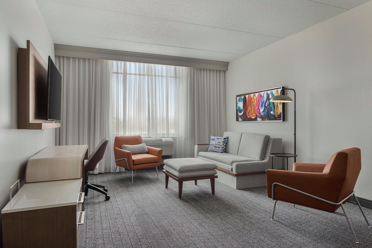 A hotel room with a desk, two armchairs, a sofa, a flat-screen TV, a floor lamp, and a large window with curtains.