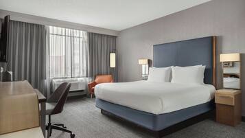 Quarto de hotel com uma cama grande, escrivaninha, cadeira, televisão e janela com cortinas.