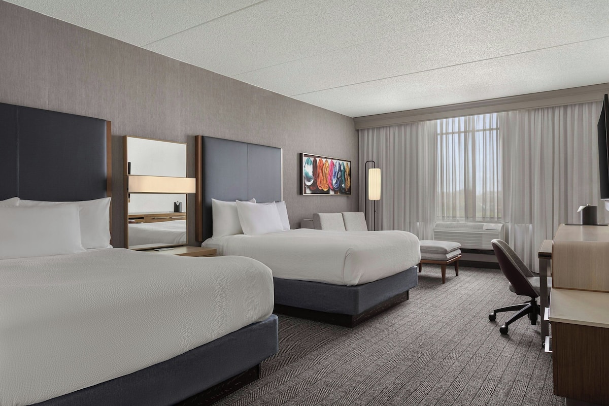 A hotel room with two beds, a desk, a chair, a lamp, and a large window with curtains.