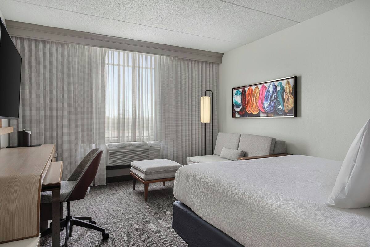 A hotel room with a bed, a desk, a chair, a lamp, and a painting on the wall.