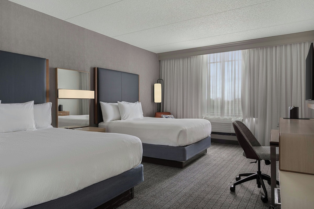 A hotel room with two beds, a desk, a chair, a large mirror, and a window with curtains.