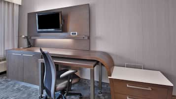 A modern hotel room with a wooden desk, a flat-screen TV mounted on the wall, a chair, and a small cabinet.