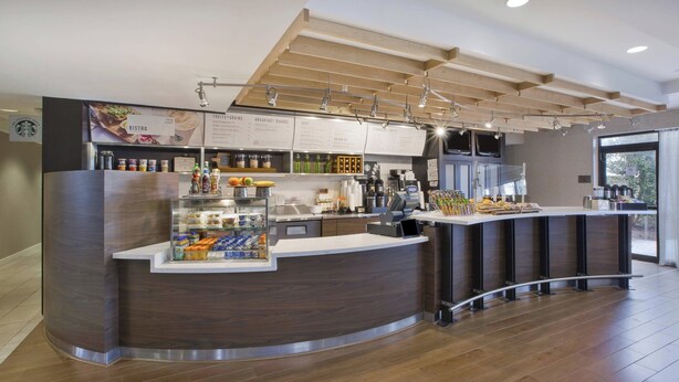 A modern bistro with a curved counter, a menu board, and a variety of food and drink items displayed. - Courtyard Somerset by Marriott (Somerset)