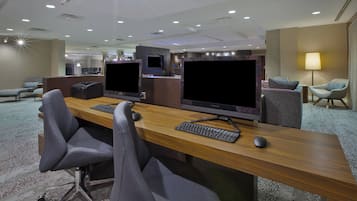 A modern hotel room with a long desk, two monitors, a keyboard, and a mouse. There are two chairs, a sofa, and a TV. The room has a carpeted floor and a ceiling with recessed lighting.
