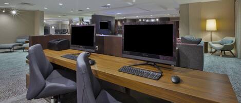 A modern hotel room with a long desk, two monitors, a keyboard, and a mouse. There are two chairs, a sofa, and a TV. The room has a carpeted floor and a ceiling with recessed lighting.
