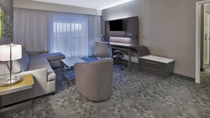 A modern hotel room with a grey sofa, a small round table, a flat-screen TV mounted on the wall, a desk with a chair, and a carpeted floor. - Courtyard Somerset by Marriott (Somerset)