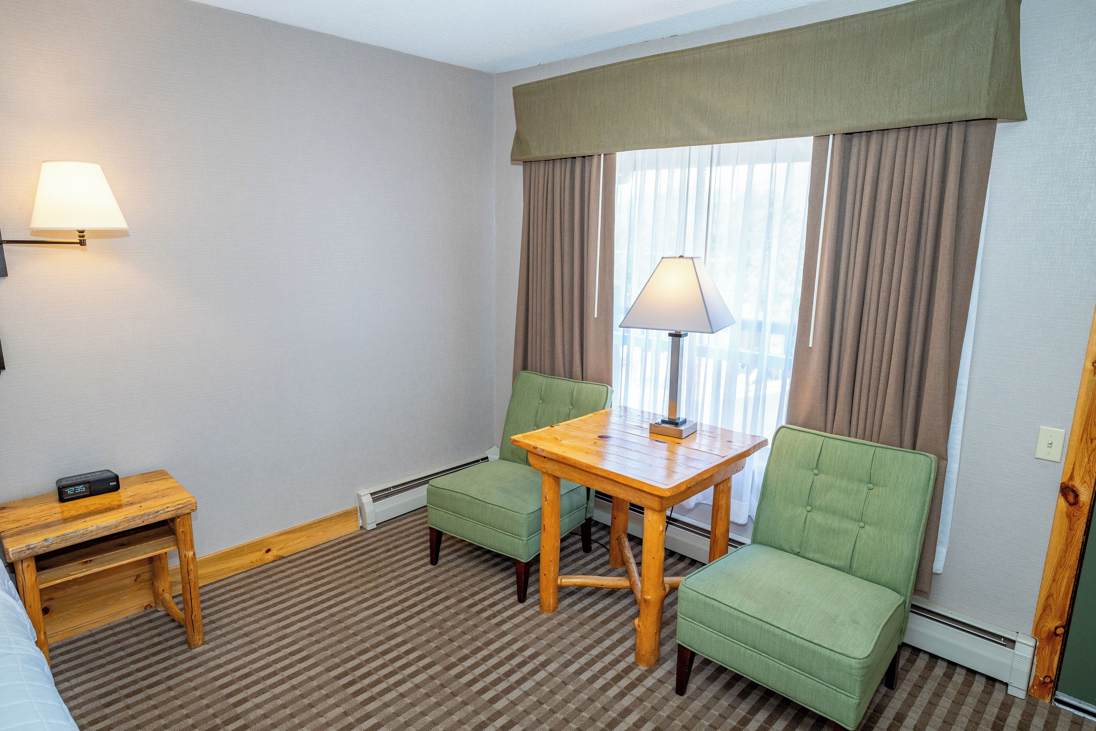 A hotel room with a checkered carpet, a wooden table with a lamp, two green armchairs, a small wooden nightstand, and a window with curtains.