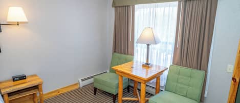 A hotel room with a checkered carpet, a wooden table with a lamp, two green armchairs, a small wooden nightstand, and a window with curtains.