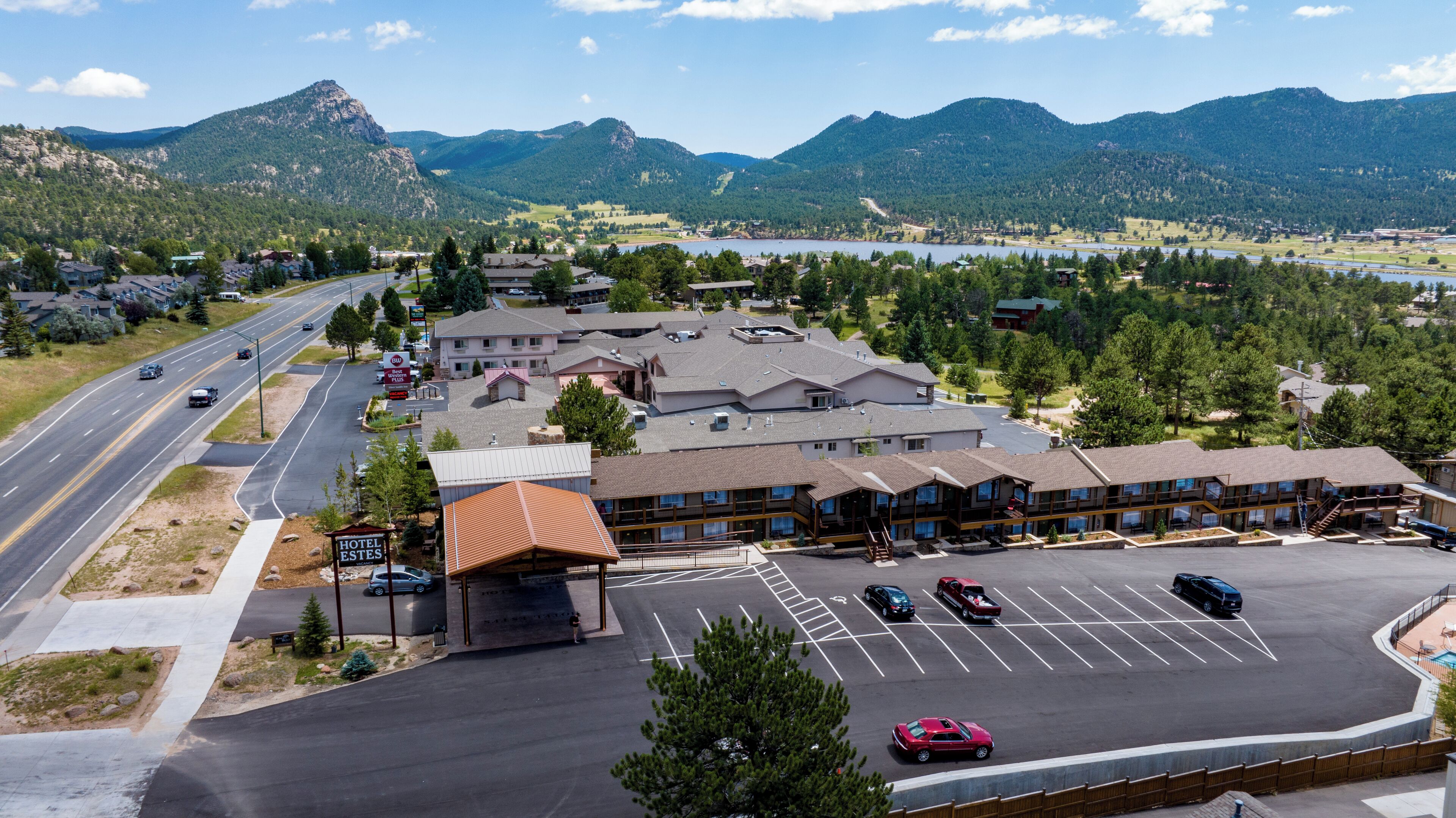 A hotel with a large parking lot, a road with vehicles, and mountains in the background.