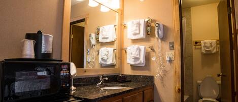A bathroom with a sink, mirror, and a microwave. There are towels hanging on the wall and a shower visible through the door.