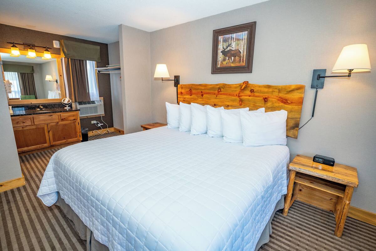 A hotel room with a large bed, a wooden headboard, a mirror, a vanity with a sink, and two bedside lamps.