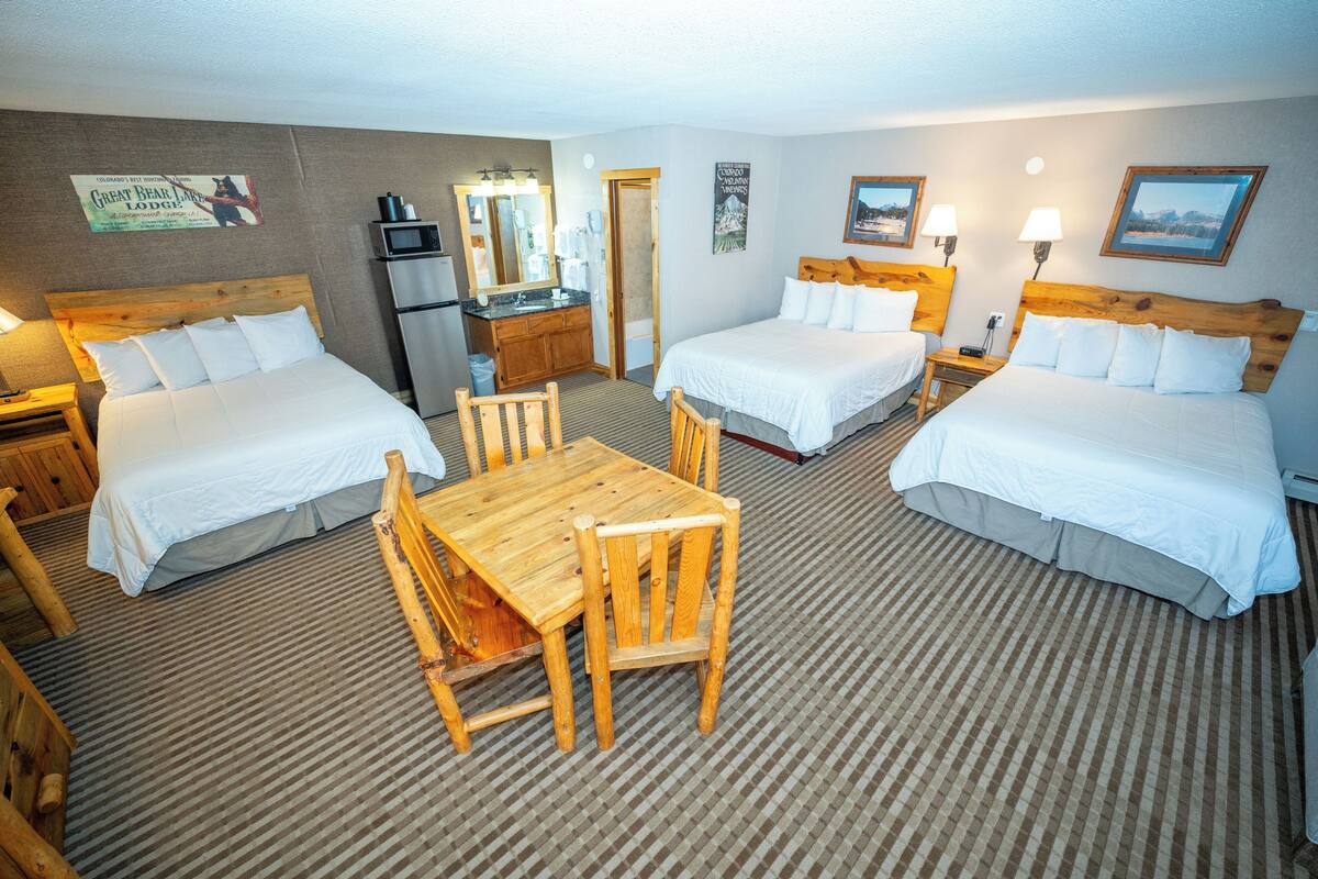 A hotel room with two beds, a wooden dining table, a kitchen area, and a bathroom.