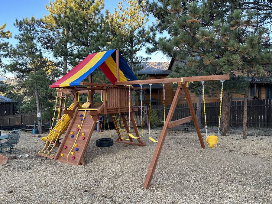 A wooden play structure with a slide and climbing wall, surrounded by trees and a wooden fence.