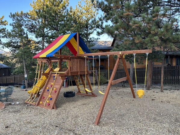 A wooden play structure with a slide and climbing wall, surrounded by trees and a wooden fence.