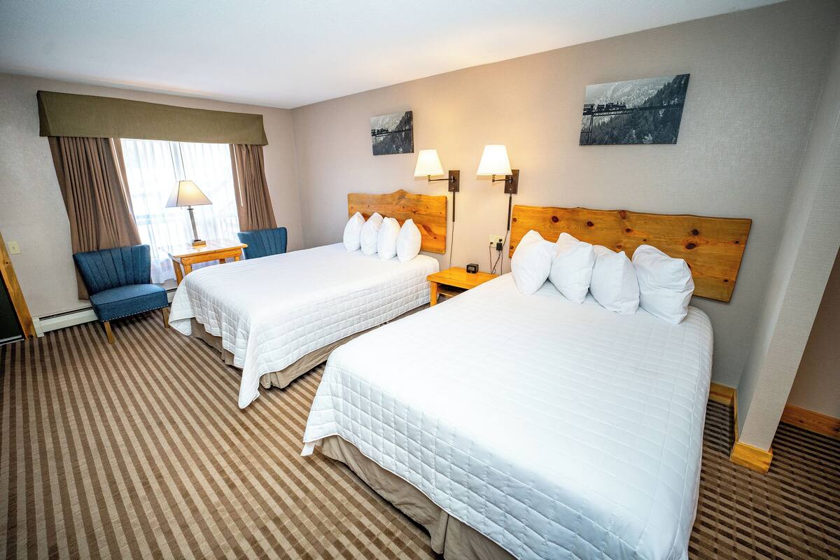 A hotel room with two beds, a chair, a table, and two wall-mounted lamps.