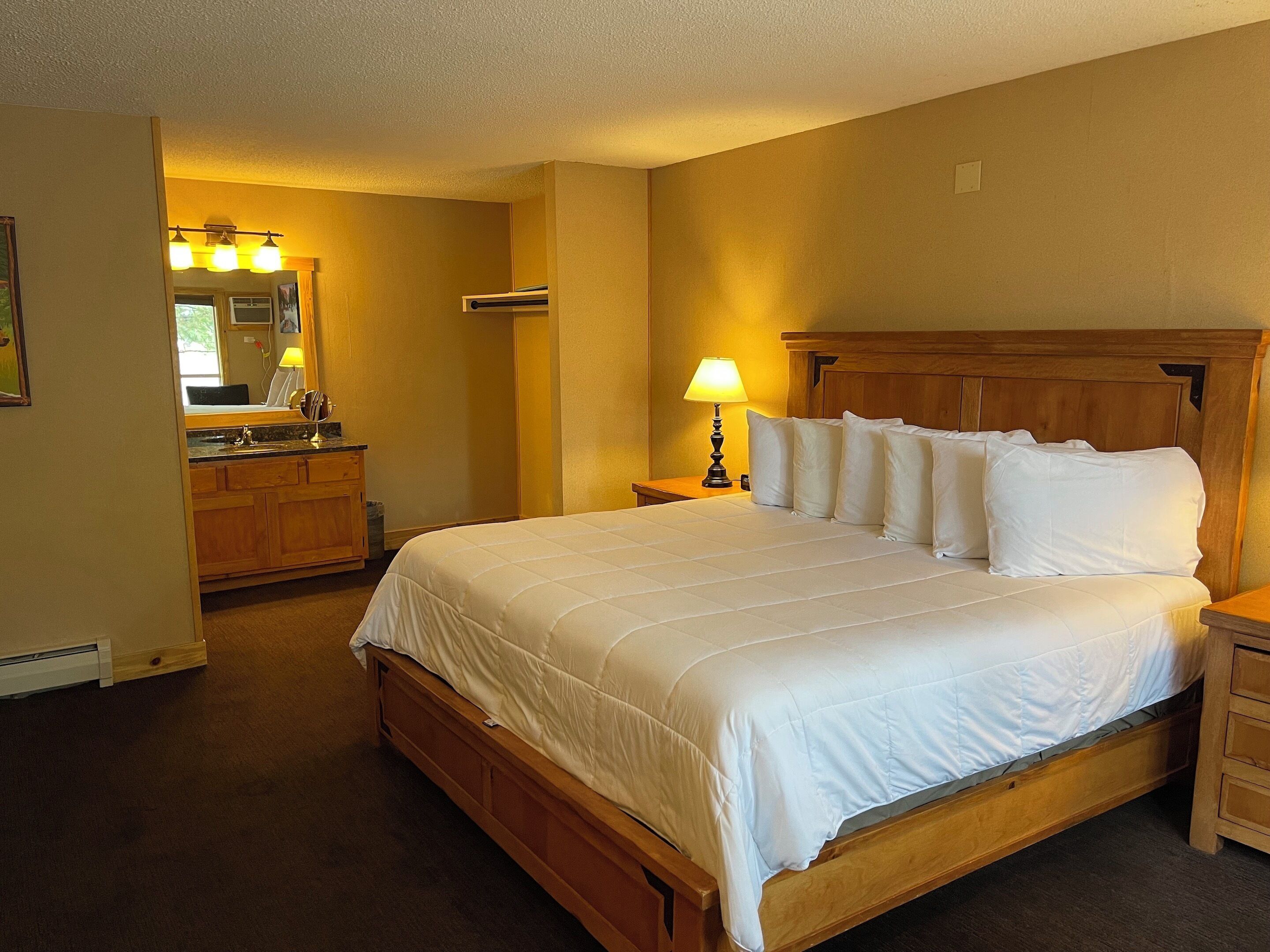 A bedroom with a wooden bed, a nightstand, a lamp, and a bathroom visible in the background.