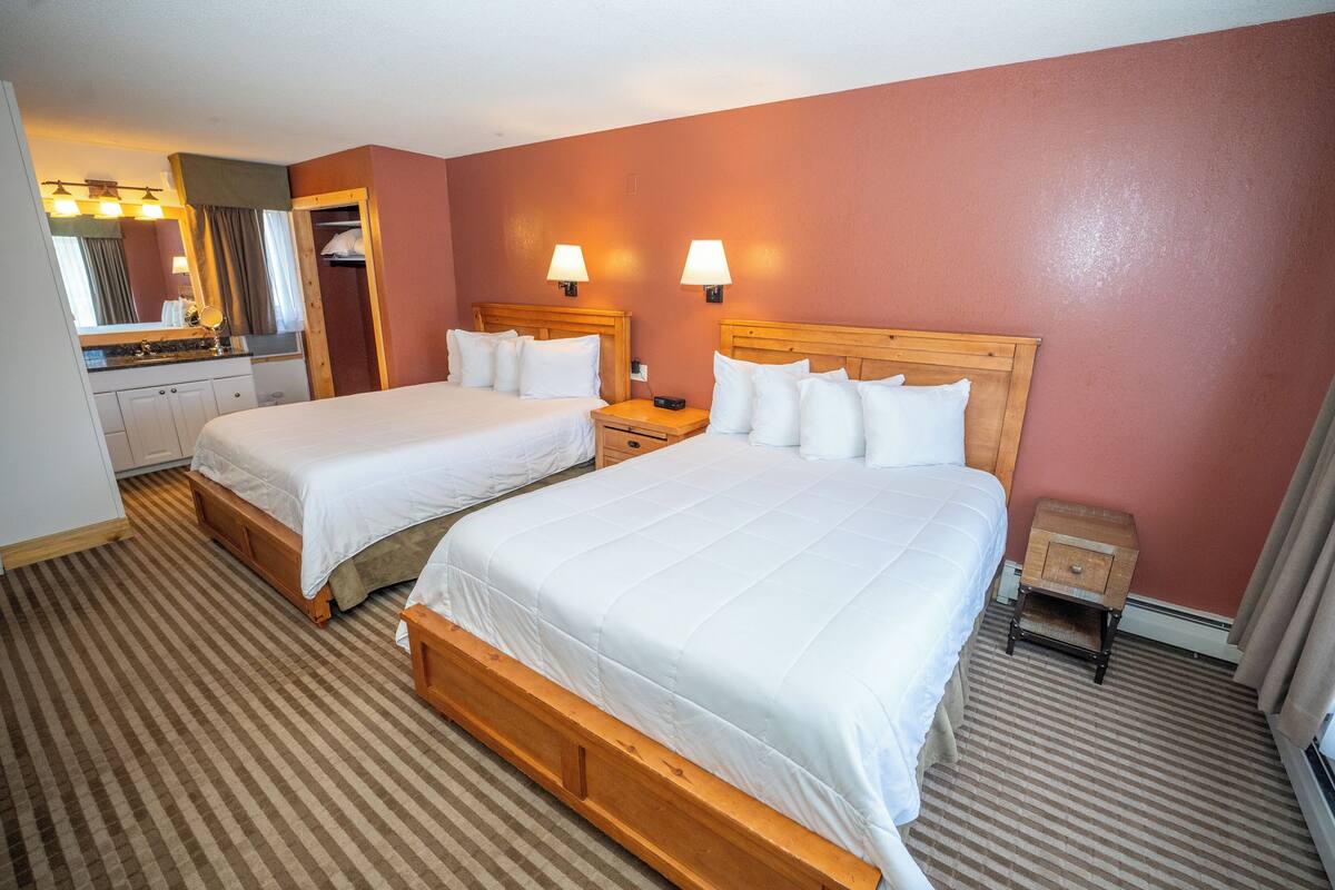 A hotel room with two beds, a wooden headboard, a small bedside table, a nightstand, a mirror, a vanity with a sink, and a striped carpet.