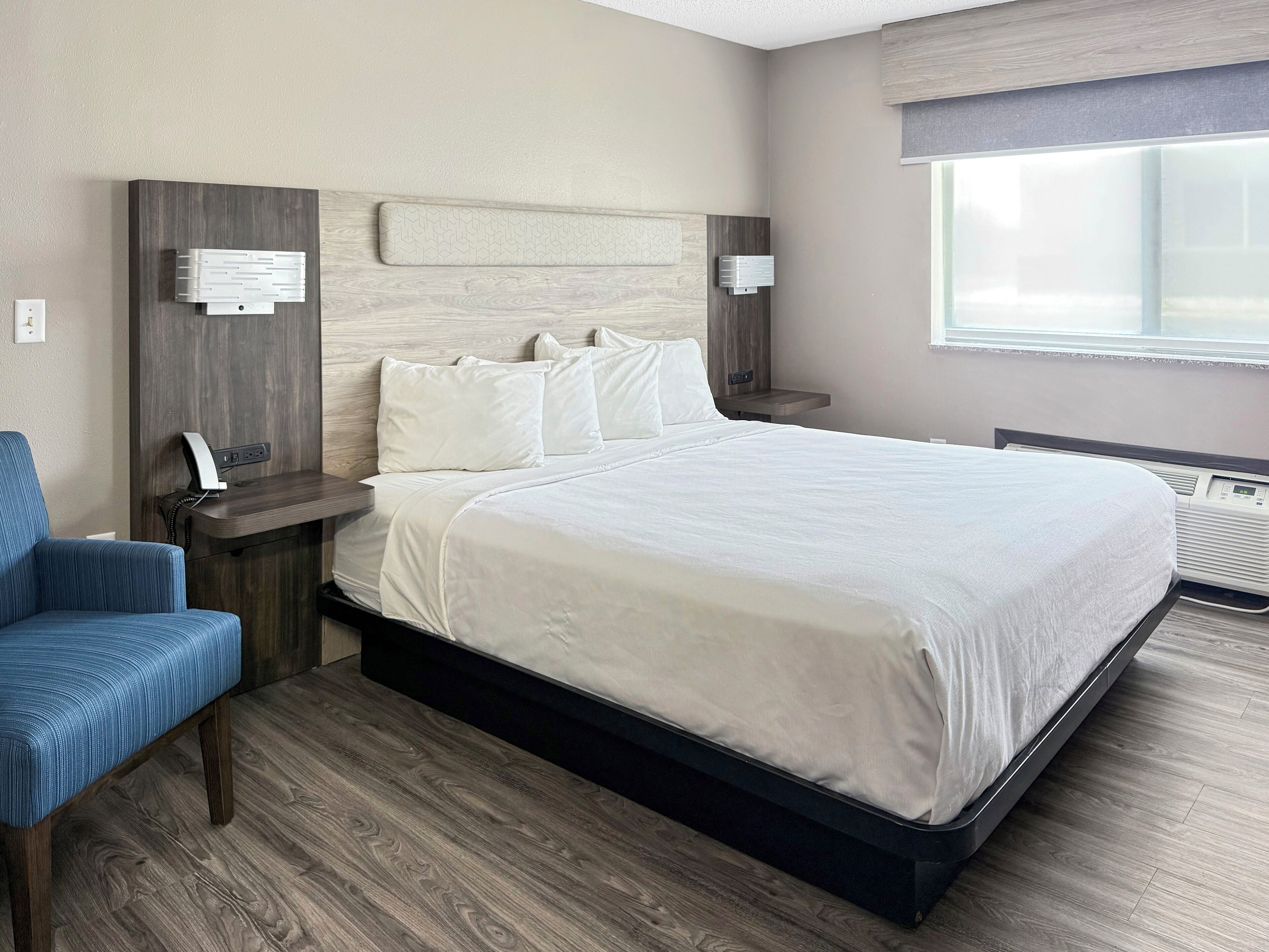 A hotel room with a large bed, a bedside table with a phone, a chair, and a window with blinds.