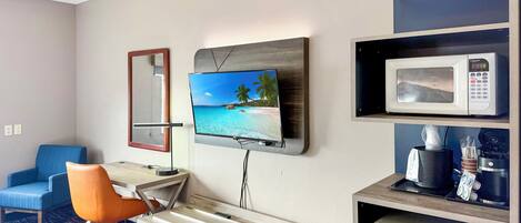 A hotel room with a TV mounted on the wall, a microwave, a small refrigerator, a desk with a chair, and a bed.
