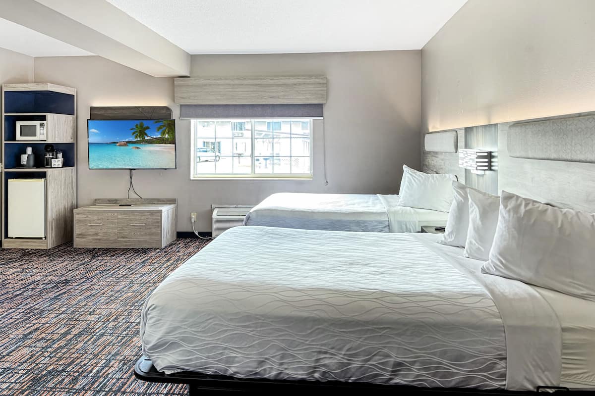 A hotel room with a bed, a TV, a desk with a microwave, and a window with a view of the beach.