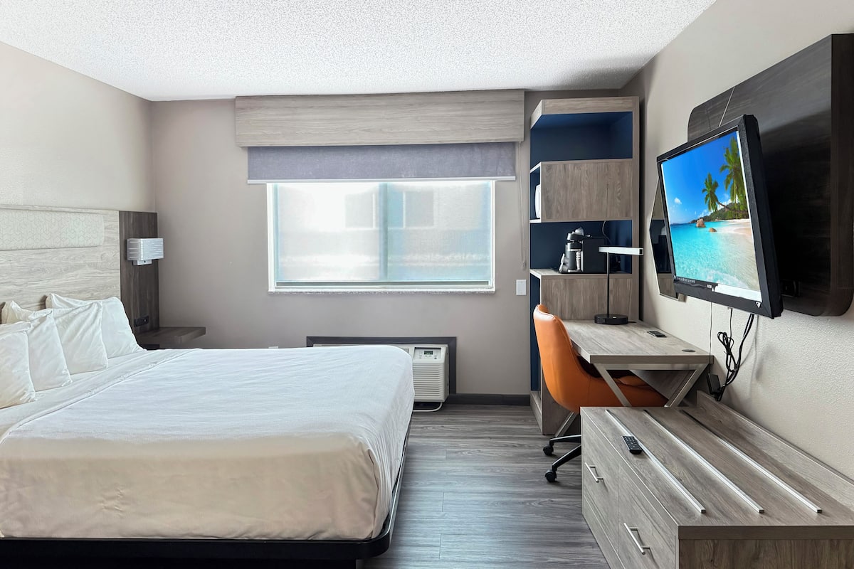 A modern hotel room with a large bed, a desk, a chair, a television, and a window with a view of the beach.