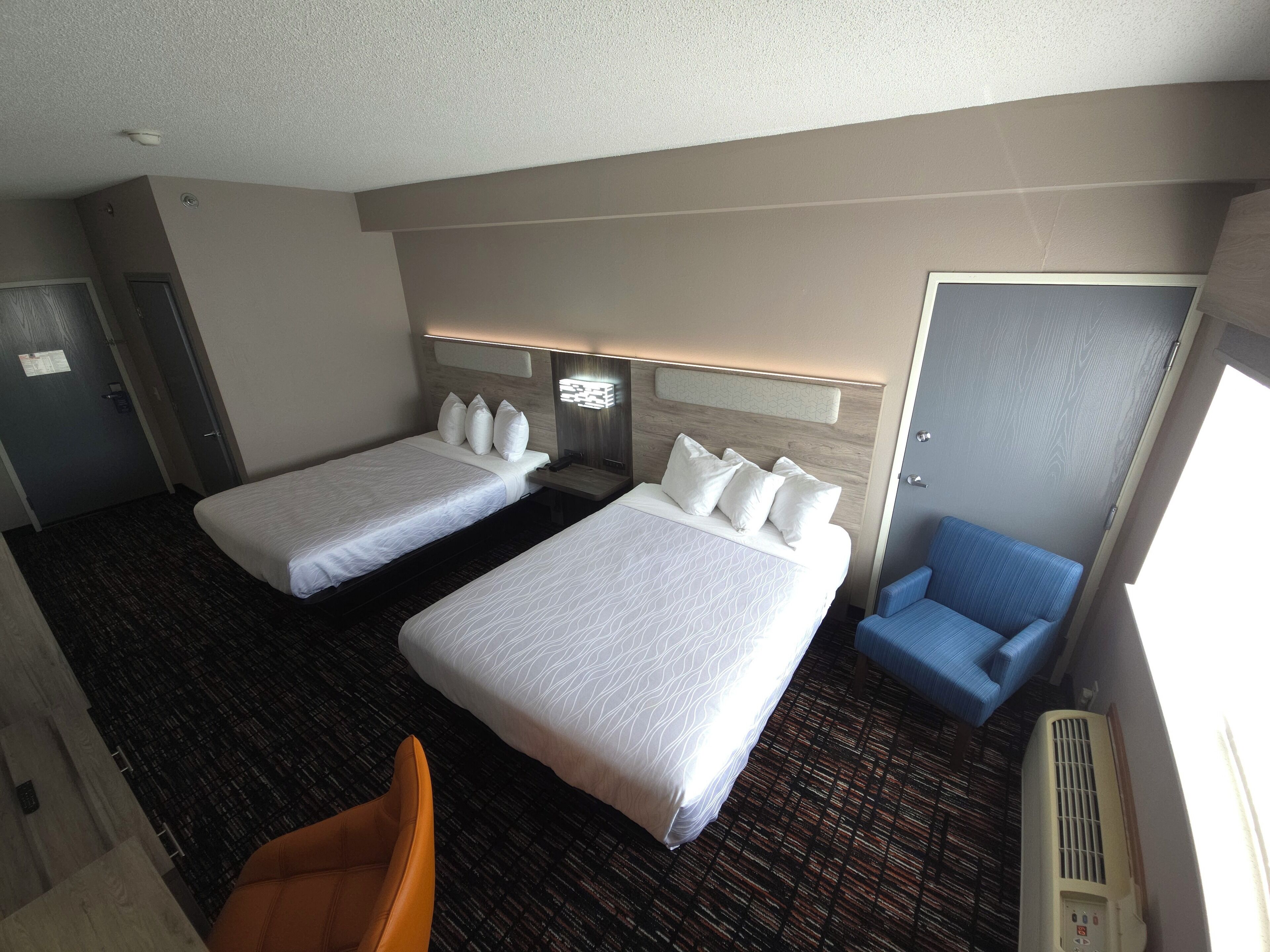 Una habitación de hotel con dos camas, un sillón azul, un escritorio con lámpara y una ventana.