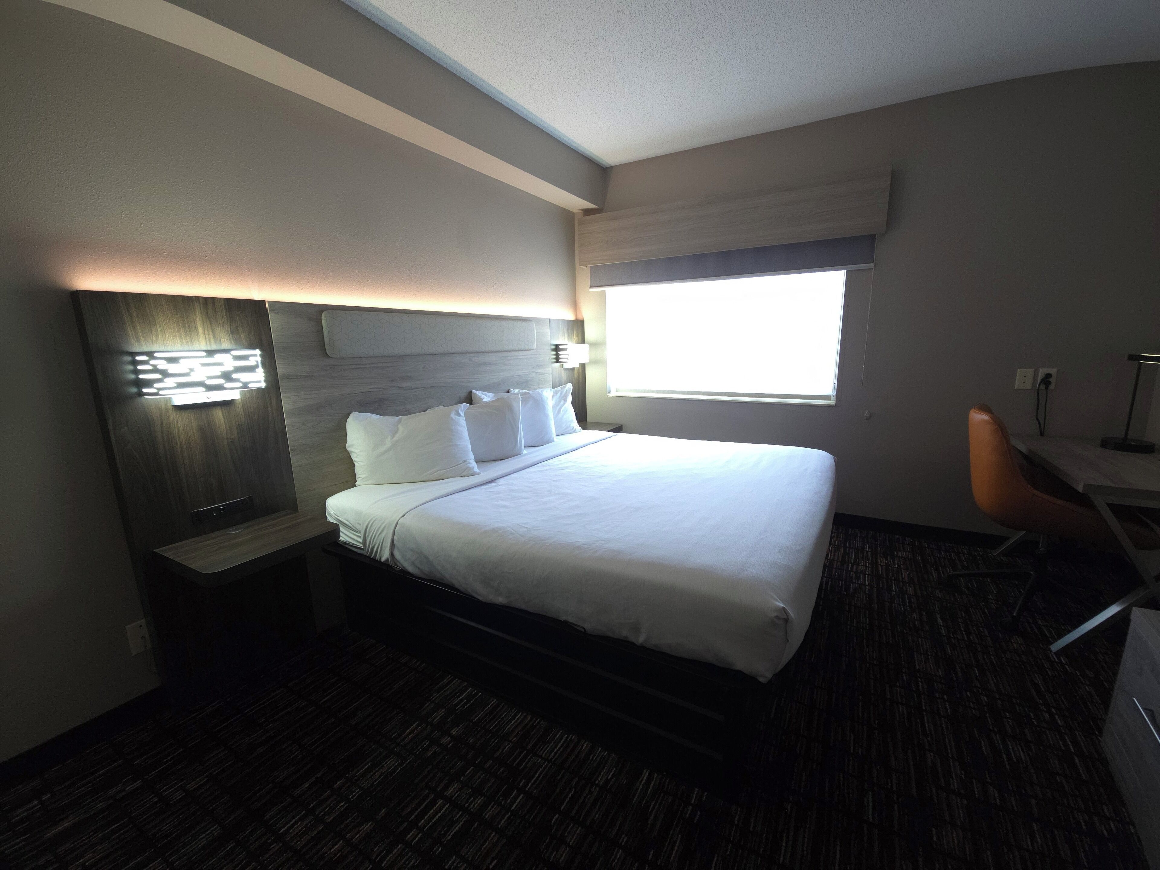 A hotel room with a bed, bedside table, lamp, and a window with curtains.