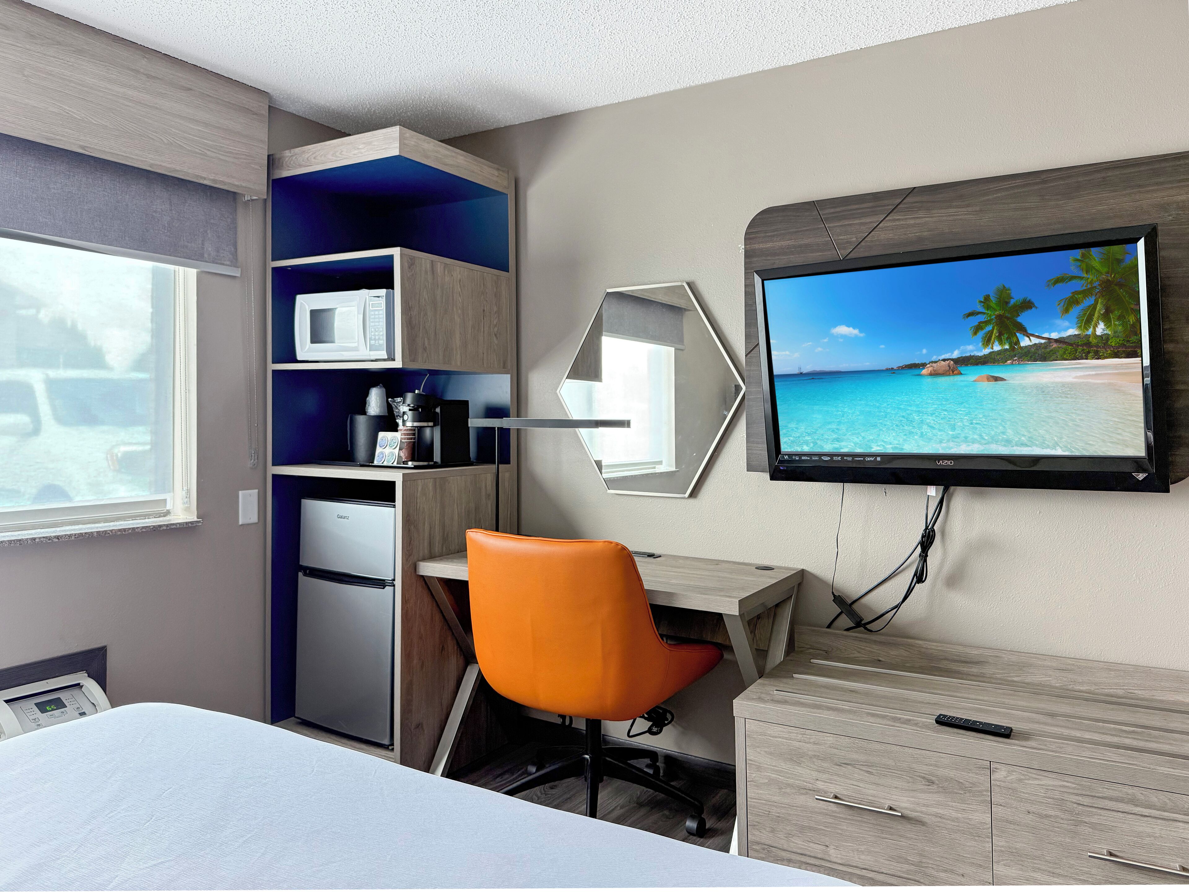 A hotel room with a bed, a desk with a chair, a TV on the wall, a microwave, a coffee maker, and a refrigerator.