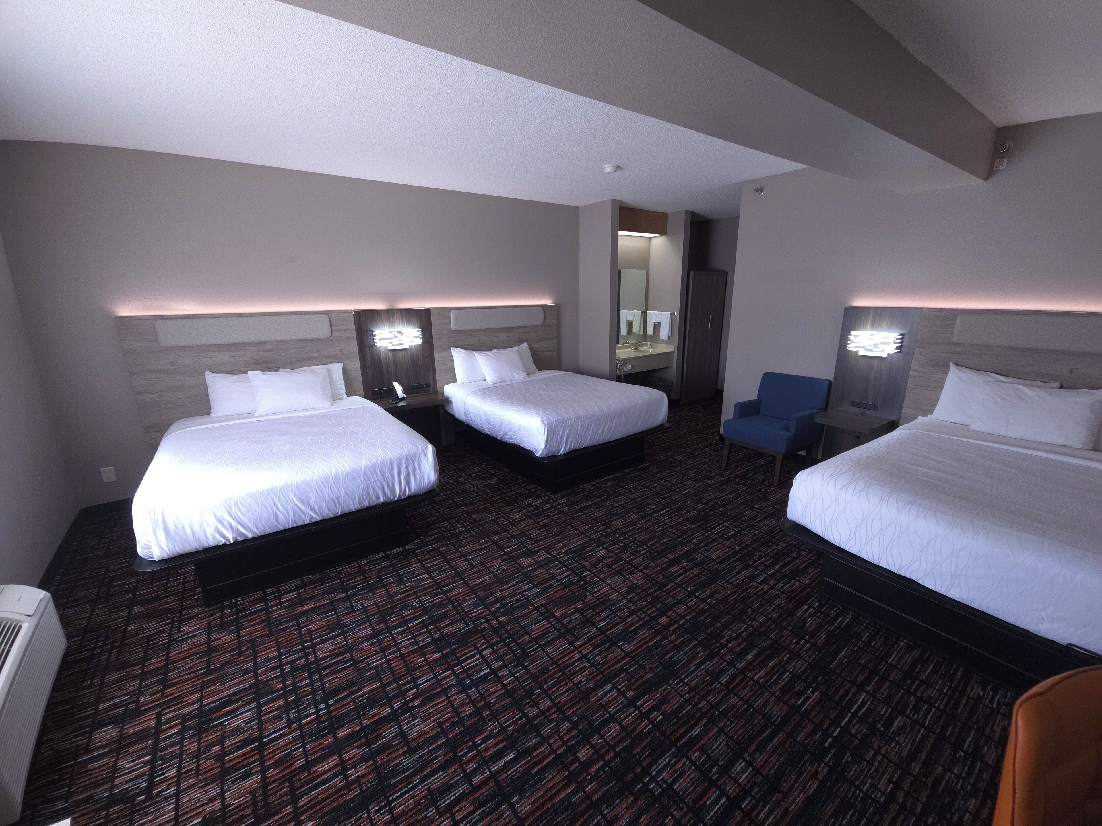 A hotel room with three beds, a small seating area, and a bathroom with a mirror and sink.