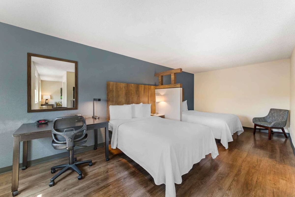 Una habitación de hotel con un escritorio de madera, dos camas, una silla y un espejo.