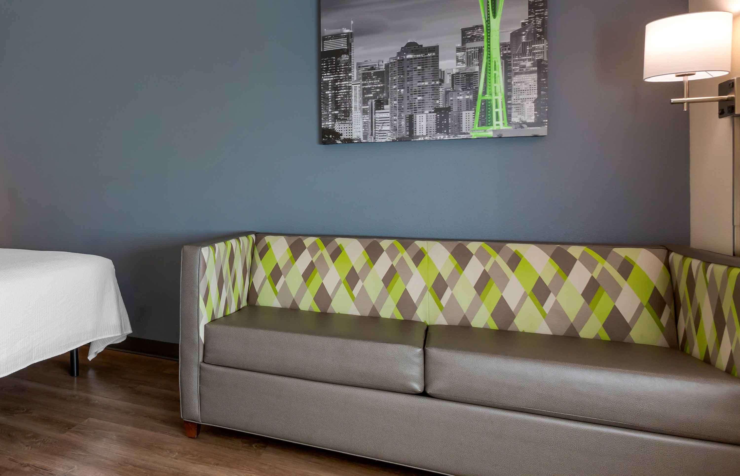 A hotel room with a grey sofa, a white bed, and a cityscape painting on the wall.
