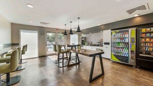 A modern restaurant with a long wooden table and chairs, a kitchen area, and vending machines.
