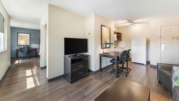 A modern hotel room with a flat-screen TV, a desk with a chair, a sofa, a refrigerator, and a mirror.
