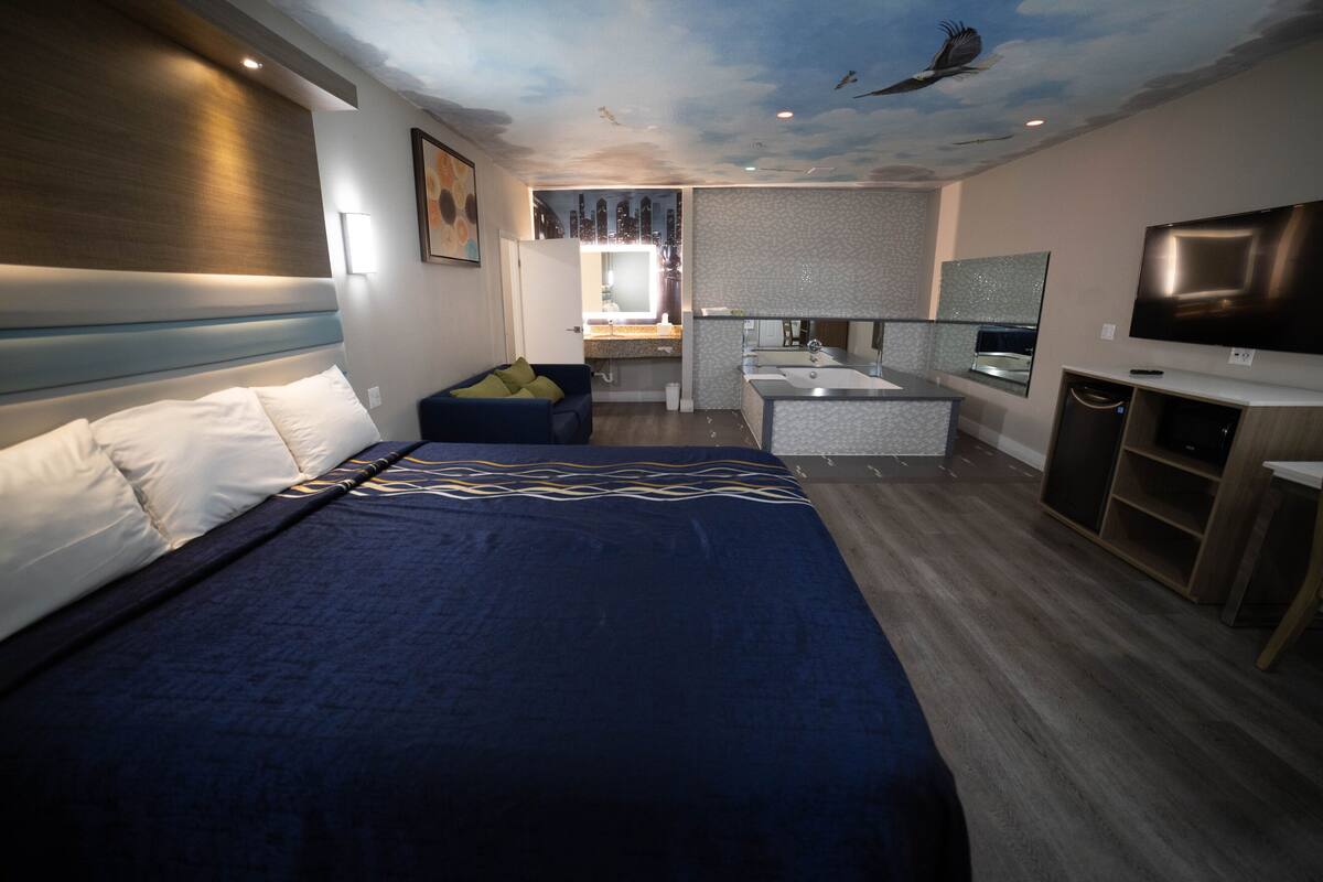 A modern hotel room with a large bed, a seating area, a bathroom with a vanity, and a television.