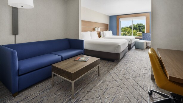 A hotel room with a blue sofa, a wooden coffee table, a bed with white bedding, and a view of greenery through a large window. - Holiday Inn Express Rochester NE- Irondequoit by IHG (Rochester)