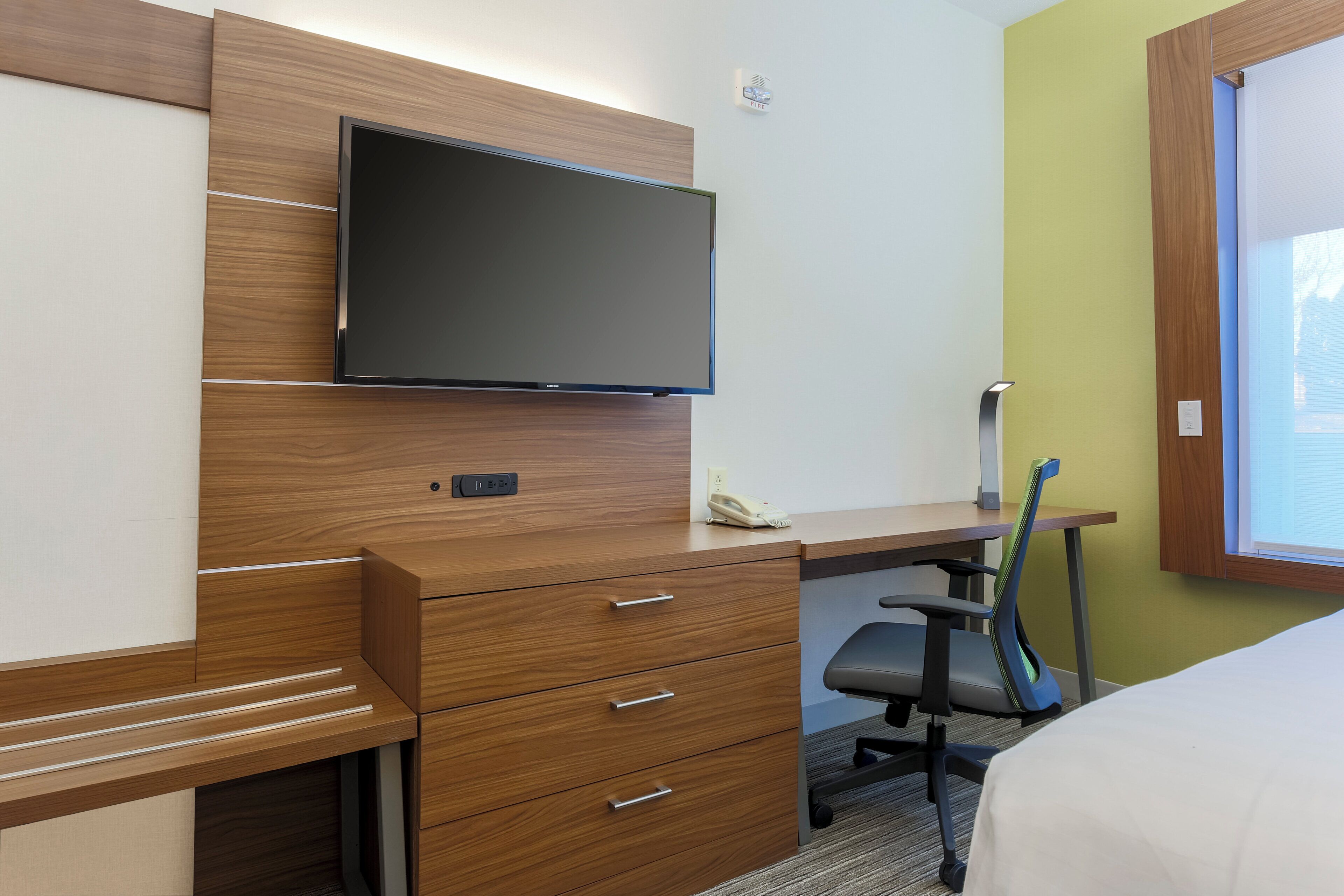 A hotel room with a wooden desk, a flat-screen TV, a telephone, a chair, and a bed.
