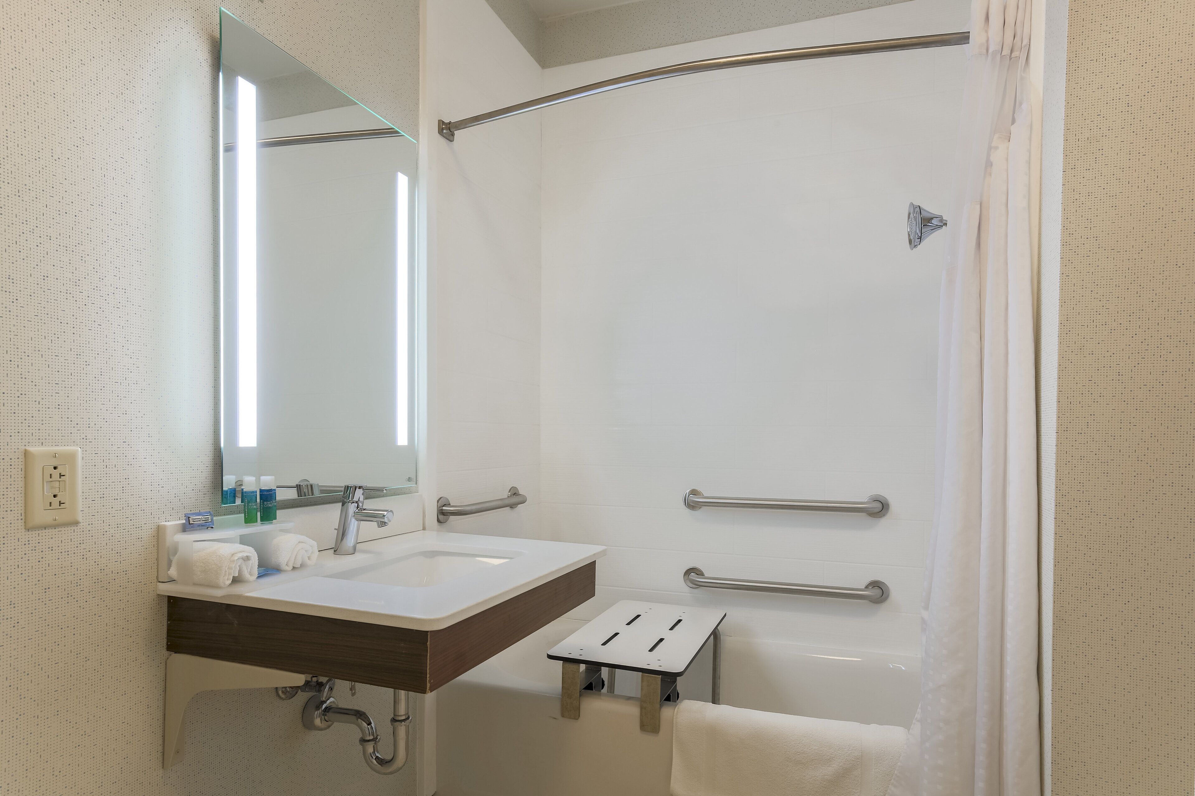 Une salle de bain avec un lavabo blanc, un miroir et une douche. On y trouve des barres d’appui et un porte-serviettes.