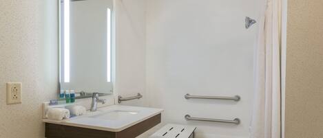 Une salle de bain avec un lavabo blanc, un miroir et une douche. On y trouve des barres d’appui et un porte-serviettes.