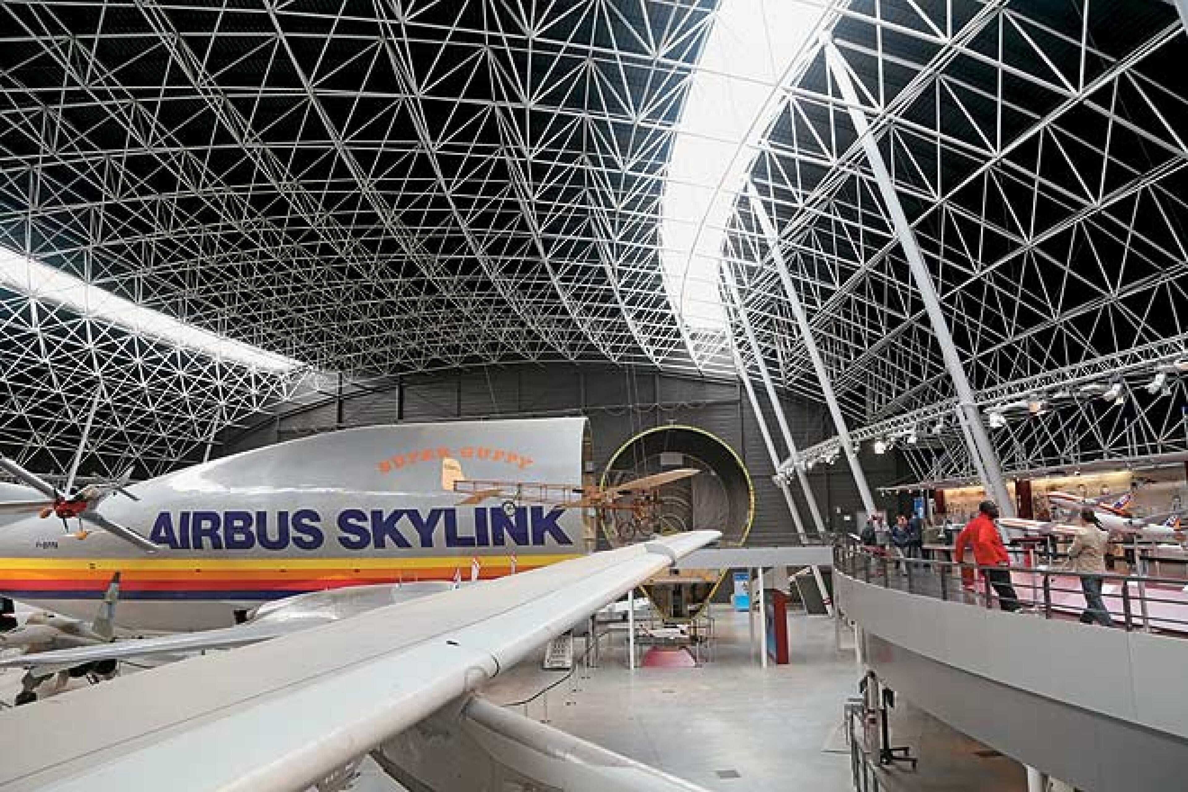 An indoor exhibition space with a large aircraft model labeled 'AIRBUS SKYLINK' and a complex metal framework ceiling.