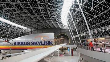 An indoor exhibition space with a large aircraft model labeled 'AIRBUS SKYLINK' and a complex metal framework ceiling.
