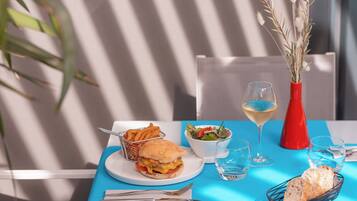 Une table avec un hamburger, une salade et des boissons, dressée sur une nappe bleue, agrémentée d’un vase rouge et d’une plante.