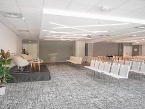 A modern conference room with a carpeted floor, white chairs, a podium, and a sofa. - Holiday Inn Toulouse Airport by IHG (Blagnac)