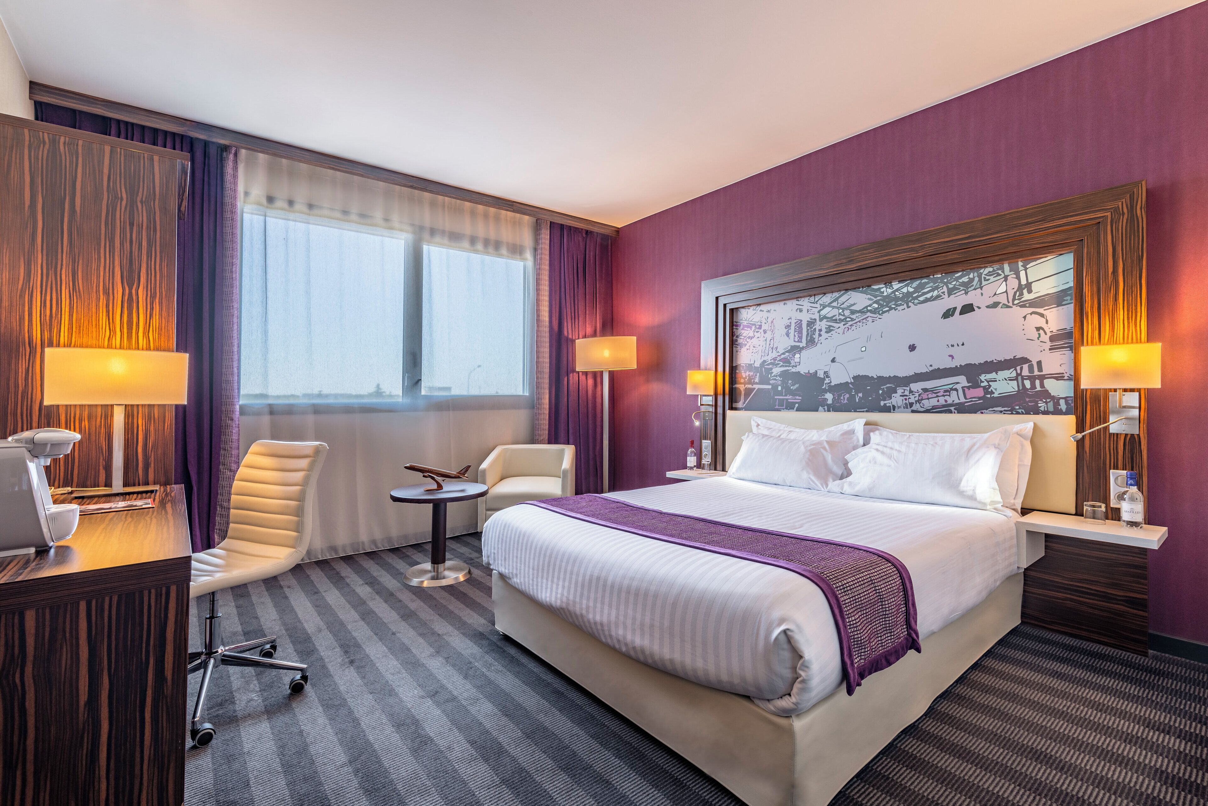 A hotel room with a bed, two chairs, a desk, and a large window.