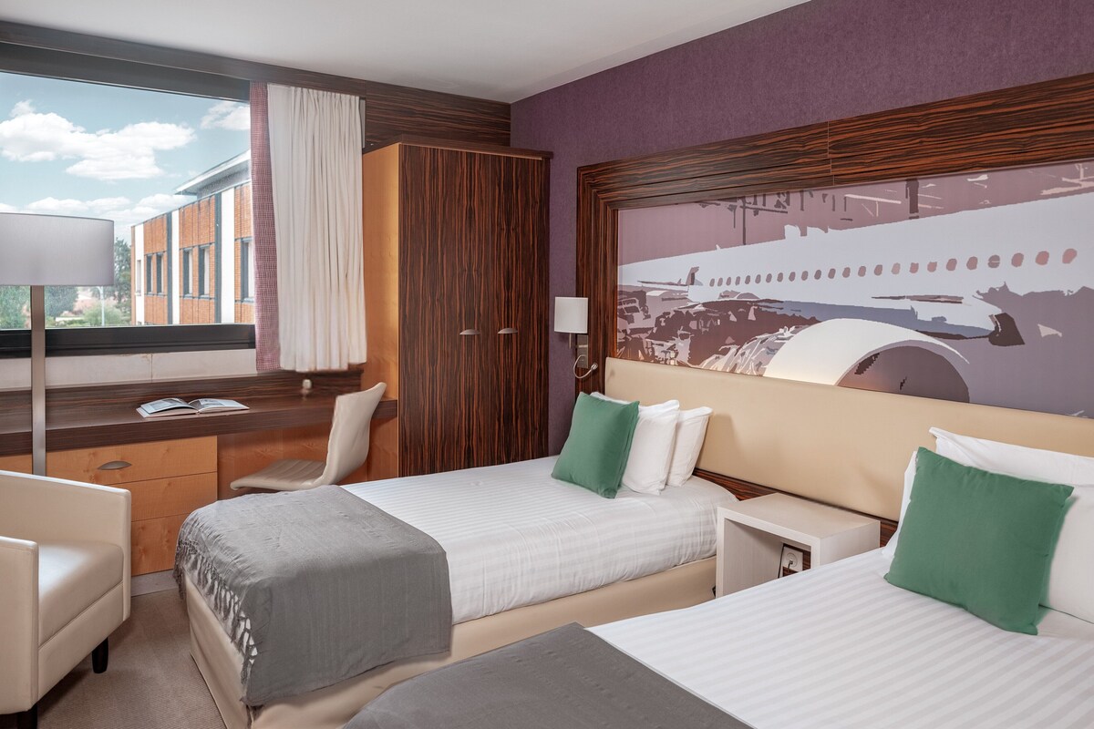 A hotel room with two beds, a desk, a chair, and a large window with a view of buildings.