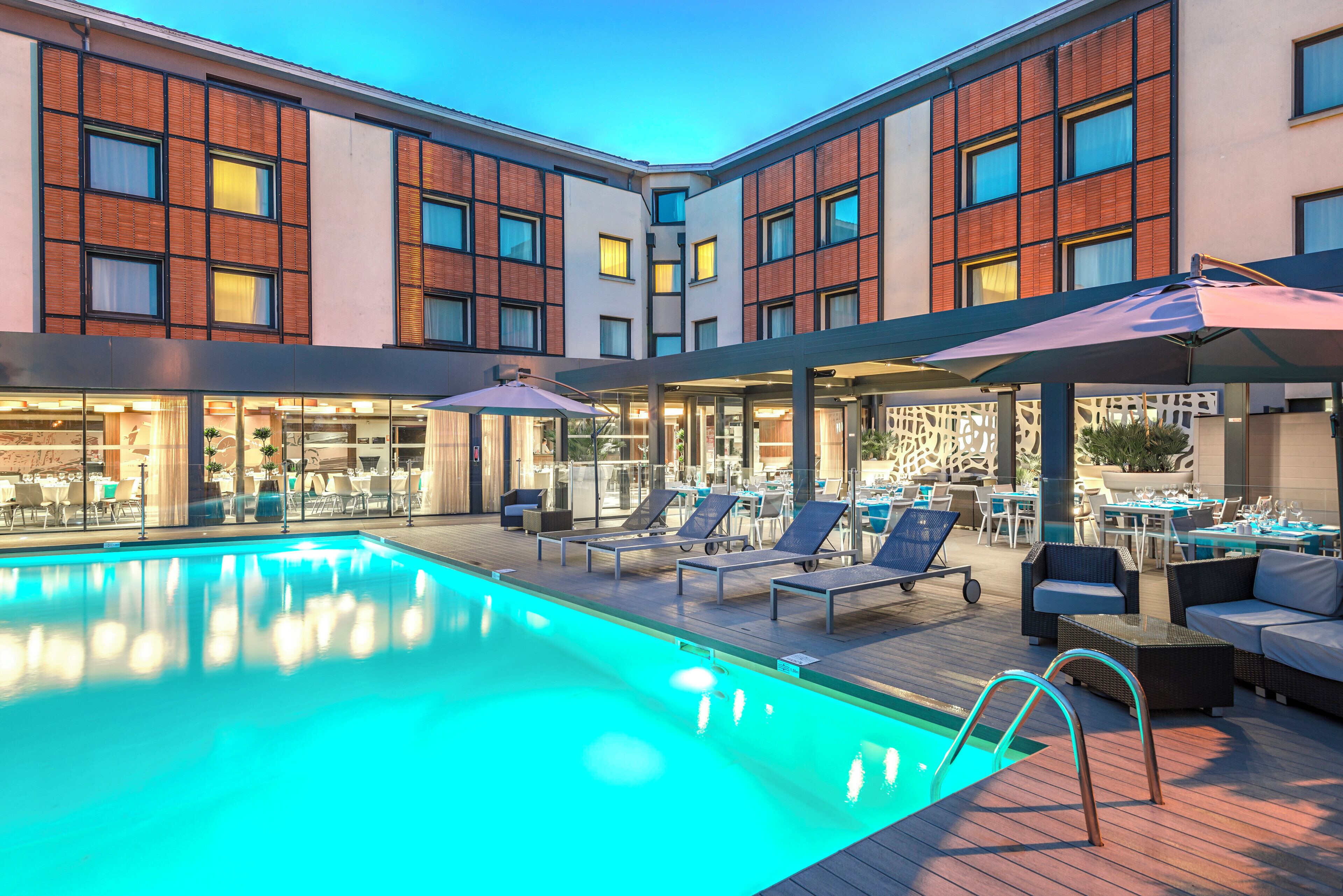 A hotel pool area with lounge chairs and tables, surrounded by a building with large windows and a brick facade.