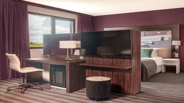 Habitación de hotel con escritorio, silla, televisor, cama y vistas a la ciudad a través de la ventana.