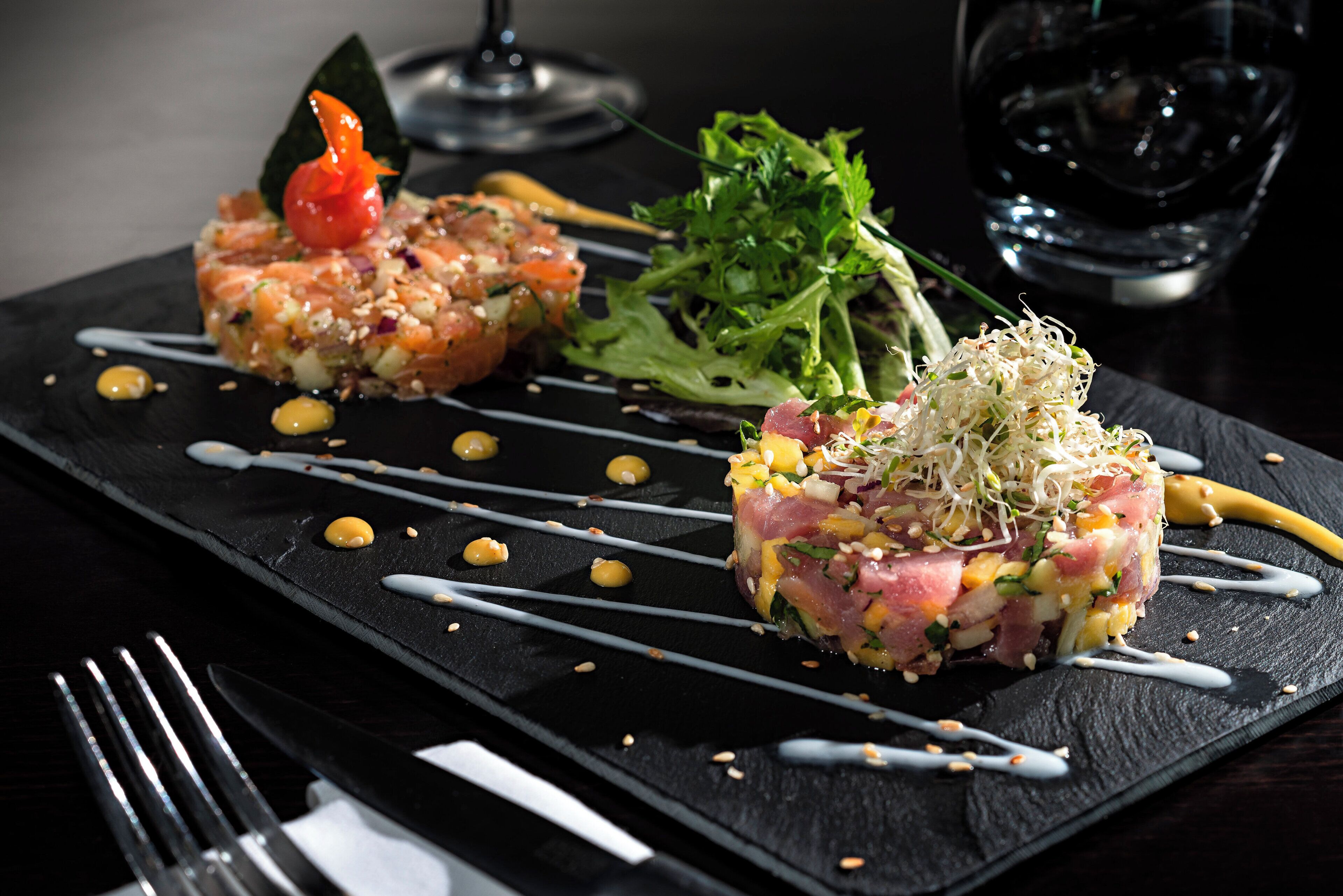 A black slate serving board with two dishes of raw fish salad, garnished with seeds and a small red vegetable, accompanied by a green salad and a glass of red wine.