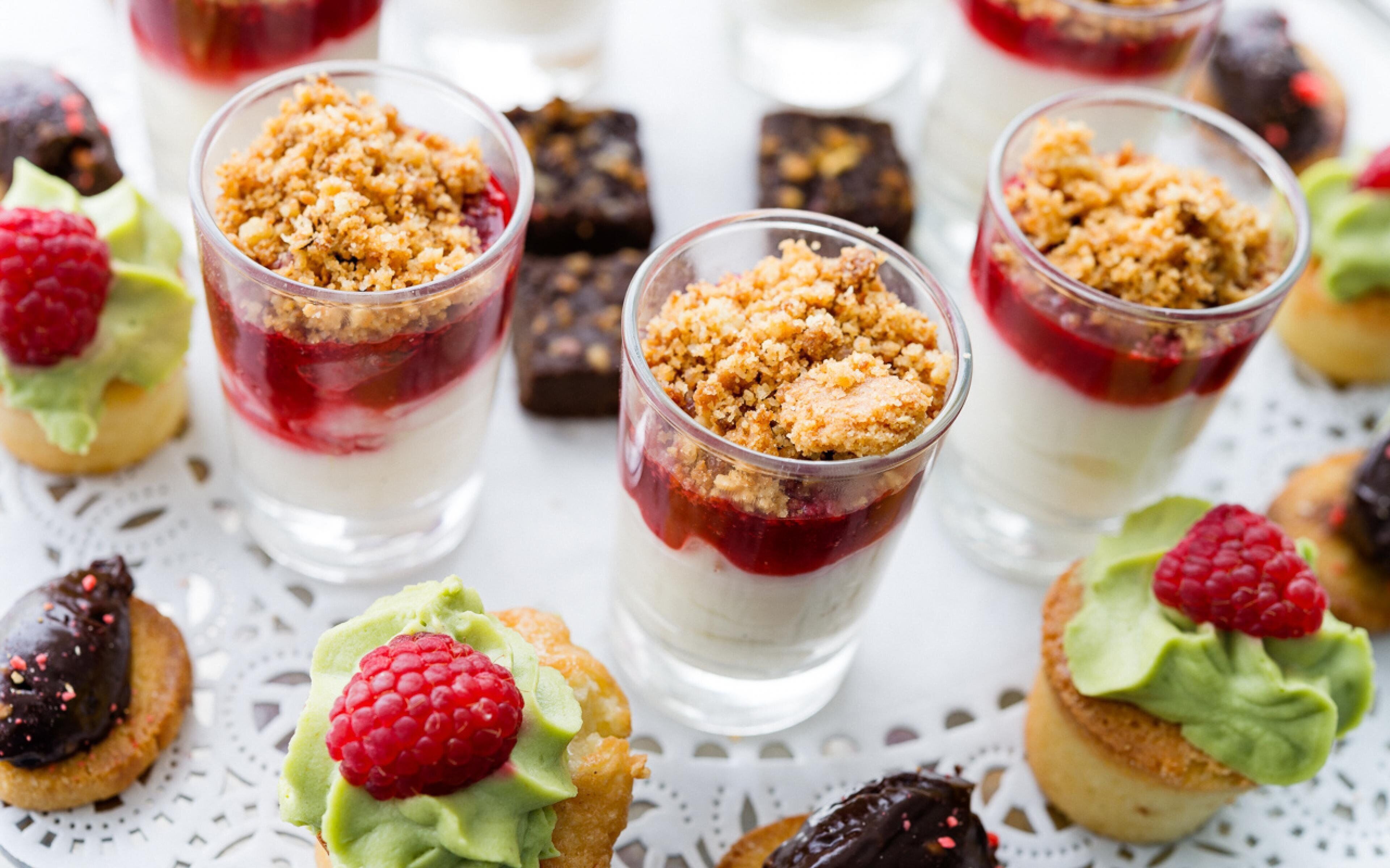 Une présentation de desserts comprenant des couches de différentes sortes de desserts dans des verres transparents, le tout surmonté de miettes de pâtisserie et décoré de framboises.
