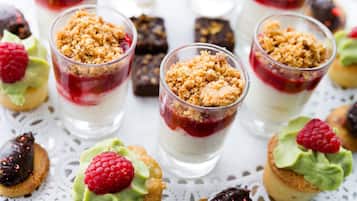 Une présentation de desserts comprenant des couches de différentes sortes de desserts dans des verres transparents, le tout surmonté de miettes de pâtisserie et décoré de framboises.