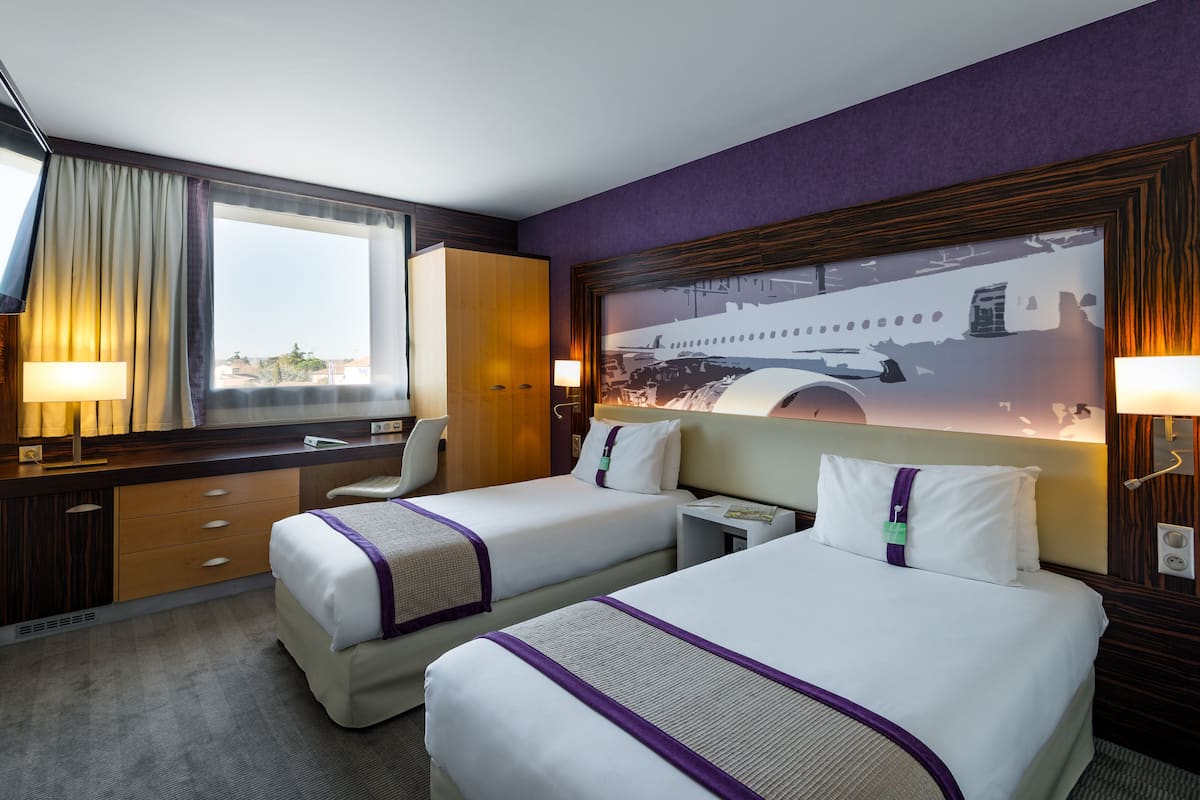 Ein Hotelzimmer mit zwei Betten, einem Schreibtisch, einem Stuhl, einem Fenster mit Vorhängen und einer Wandlampe.